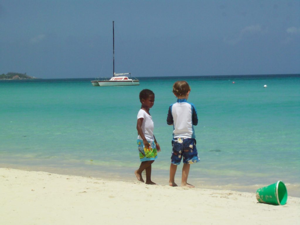 toddlers on white sand beach with turquoise water and sailboat in background